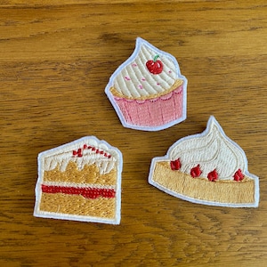 May include: Three embroidered patches depicting desserts. One is a cupcake with pink frosting, white icing, and a cherry. Another is a slice of cake with red filling and white frosting. The third is a slice of cake with white frosting and red berries.