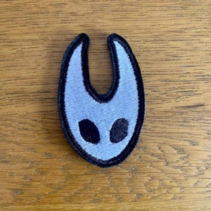 May include: A light blue embroidered patch with a mask-like design and two black eye shapes. The patch has a black border and is on a wooden surface. This patch is suitable for clothing or accessories. The design is stylized.