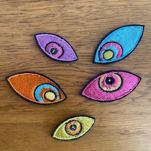 May include: Five embroidered eye-shaped patches with black borders. The top left is purple with a pink iris, the top right is blue with a pink and yellow iris, the middle left is orange with a blue and yellow iris, the middle right is pink with a yellow iris, and the bottom is yellow with an orange iris.
