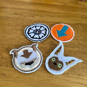 May include: Four embroidered patches featuring characters from the animated television series Avatar: The Last Airbender. The patches are white with brown, blue, and orange accents. The patches feature the symbols of the four elements: water, earth, fire, and air.