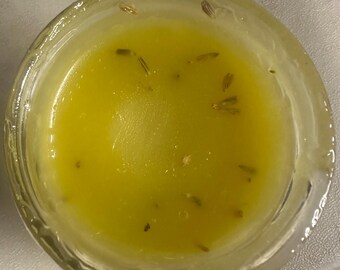 May include: A clear glass jar filled with a vibrant yellow liquid, possibly oil or a sauce. Small, dark specks, likely herbs or spices, are scattered throughout the liquid. The jar is sitting on a light-colored surface.