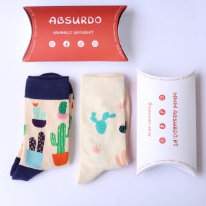 Pode incluir: Dois pares de meias com estampa de cacto. Um par é branco com um cacto verde e o outro par é azul marinho com uma estampa de cacto multicolorido. As meias são embaladas em uma caixa de travesseiro branca com o texto "WWW.ABSURDO.PT" e as alças de mídia social para Instagram, TikTok e Facebook.