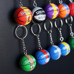 May include: A collection of eight basketball keychains featuring different team logos and colours. The keychains are made of plastic and feature a basketball design with a metal ring.