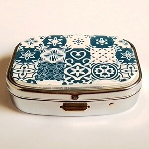 May include: A small, rectangular pill box with a silver metal frame. The lid features a patchwork design of teal and white geometric patterns. The box is closed, with a small latch visible on the front.