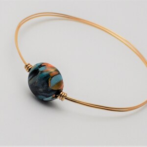 May include: A gold wire bangle bracelet with a single round bead. The bead is made of black, blue and orange marbled clay.