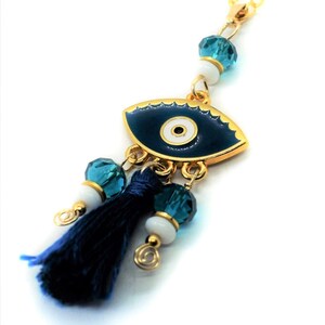 May include: A gold-colored chain with a dark blue enamel eye pendant. The pendant has a gold border and a white center. It is adorned with blue crystal beads, white beads, and a dark blue tassel. The eye charm is a symbol of protection.