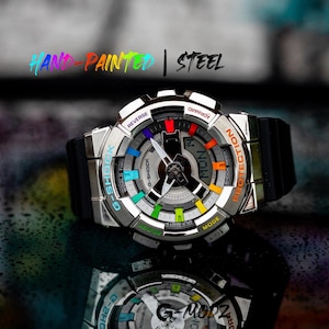 Puede incluir: Un reloj G-Shock plateado y negro con una esfera de colores arcoíris y una correa de goma negra. La esfera del reloj muestra el día y la fecha. El texto "HAND-PAINTED | STEEL" está encima del reloj.