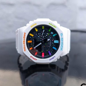 Puede incluir: Reloj G-Shock blanco con esfera negra y detalles coloridos. El reloj presenta una pantalla digital con marcadores de colores del arcoíris y texto que dice "PROTECTION", "START", "LIGHT" y "ADJUST". El reloj está sobre una superficie reflectante.