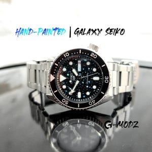 May include: A hand-painted Galaxy Seiko watch with a stainless steel band. The watch face features a black background with a galaxy design. The bezel is black with white markings. The text "HAND-PAINTED | GALAXY SEIKO" is above the watch.