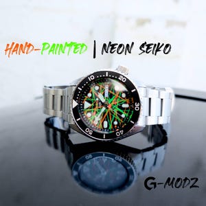 May include: A stainless steel wristwatch with a black bezel and a dial featuring neon green and orange accents. The watch has a silver band and is reflected on a glossy black surface. Text on the watch reads "HAND-PAINTED | NEON SEIKO".