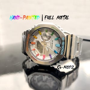 Puede incluir: Un reloj G-Shock plateado con esfera blanca y detalles coloridos. El reloj presenta el texto "HAND-PAINTED | FULL METAL" y "G-MODZ". Tiene una pantalla digital que muestra la hora.