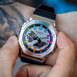 May include: A silver G-Shock watch with a black band and a rainbow colored face. The watch face has a digital display showing the time and date. The watch has buttons labeled "Adjust", "Mode", "Light", "Start", and "Protection".