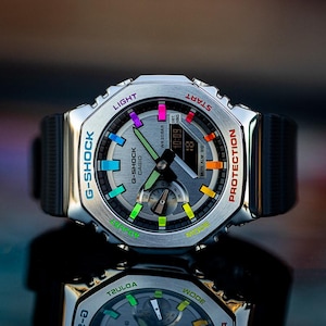 May include: A silver G-Shock watch with a black band. The watch face has a white background with a multi-colored ring around the edge. The ring has the words "G-Shock", "Casio", "Light", "Start", "Mode", "Protection", and "Tzuloa" printed on it.