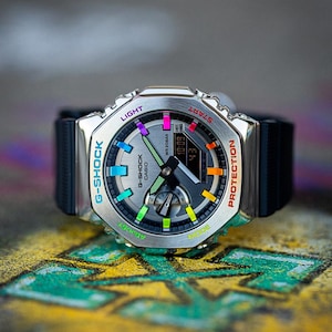 May include: A silver G-Shock watch with a black band. The watch face has a rainbow colored design with the words "G-Shock" and "Casio" printed on it. The watch also has buttons labeled "Light", "Start", "Mode", "Adjust", and "Protection".