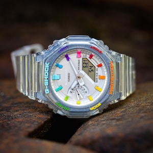 Puede incluir: Un reloj G-Shock transparente con una esfera blanca y detalles coloridos. El reloj tiene una pantalla digital con la hora, la fecha y otras funciones. El reloj está hecho de resina y tiene una resistencia al agua de 20 bares.