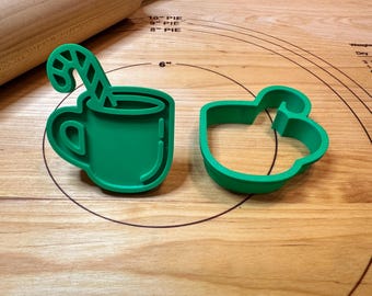 Pode incluir: Dois cortadores de biscoitos de plástico verde. Um tem a forma de uma caneca com um bastão de doces, e o outro tem a forma de uma caneca. Os cortadores estão em uma superfície de madeira com um rolo no fundo. Ferramentas de cozinha.
