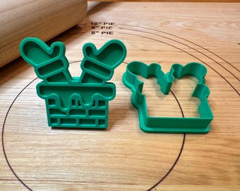 Pode incluir: Dois cortadores de biscoitos de plástico verde. Um tem a forma de uma chaminé com luvas, e o outro tem a forma de uma luva. Os cortadores estão sobre uma superfície de madeira com um rolo no fundo.