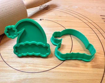 Pode incluir: Dois cortadores de biscoitos de plástico verde em forma de chapéu de Papai Noel. Um é um chapéu completo com um pompom e uma borda ondulada. O outro é o contorno de um chapéu de Papai Noel. Em uma superfície de madeira com um rolo.
