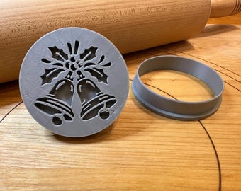 Pode incluir: Um carimbo de biscoito cinza, circular, com um desenho de azevinho e sinos, ao lado de um anel de cortador de biscoito cinza correspondente. Um rolo de madeira no fundo. Ideal para assar nas festas.