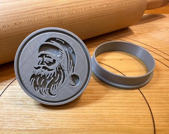 Pode incluir: Um cortador de biscoitos circular cinza com um design detalhado do rosto do Papai Noel. Ao lado, há um anel cinza separado, provavelmente para moldar a massa. Um rolo de madeira no fundo sugere assar.