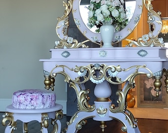 May include: A white vanity table with gold accents and a large ornate mirror. The table has a pink painted finish and features intricate carvings. A small stool with a floral patterned seat sits in front of the table.