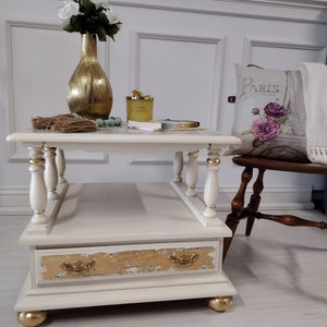 May include: A cream-colored, two-tiered coffee table with gold accents. The table features a drawer with gold leaf detailing and ornate handles. The legs are turned and painted cream with gold accents. A gold vase and other decorative items sit on top.