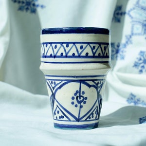 May include: A white ceramic cup with blue and white geometric designs. The cup is sitting on a white fabric with blue embroidered designs.