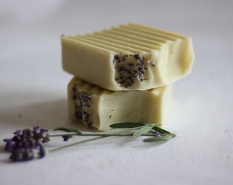 Puede incluir: Dos barras rectangulares de jabón artesanal apiladas. El jabón es de color amarillo pálido, con una parte superior texturizada y pequeños capullos de lavanda morada incrustados en los lados. Una ramita de lavanda está cerca.
