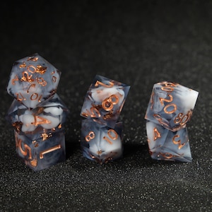 May include: A set of six polyhedral dice with a white and grey marbled effect and copper-colored numbers. The dice are arranged in a row on a black surface.