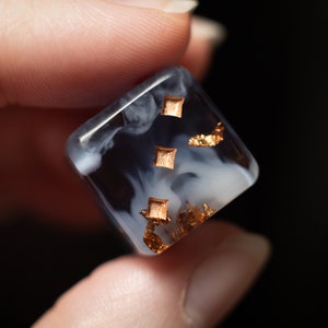 May include: A clear resin dice with white swirls and gold flecks. The dice has a square shape and is showing the number three.