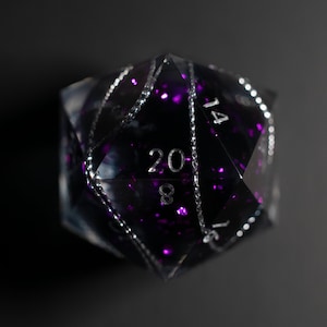 May include: A large, clear, twenty-sided die with a dark interior filled with purple glitter. The numbers are silver, and the edges are outlined with a silver chain-like design. The die is set against a dark background.