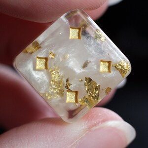 May include: A white, square-shaped dice with gold flecks and gold squares on each side.