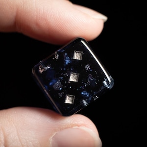 May include: A dark blue, square die with three silver pips. The die has a speckled, galaxy-like appearance with embedded silver flakes. The die is held between two fingers against a black background.