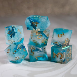 Puede incluir: Juego de siete dados poliédricos con temática marina. Los dados son transparentes con un diseño arremolinado azul y blanco, y números dorados. Los dados están apilados uno encima del otro, con un fondo borroso.