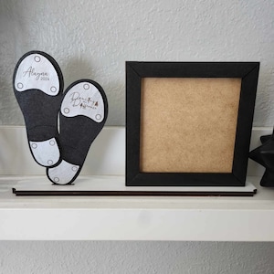 May include: Two black tap shoes with white soles and silver accents. The shoes are on a shelf with a black picture frame. The shoes have the text "Alaina 2023" and "Dancing for a Difference" on them.