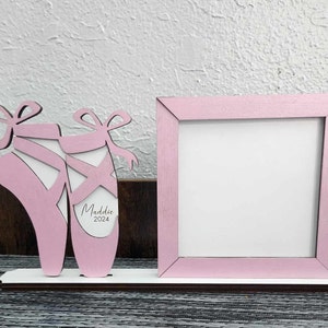 May include: Pink wooden picture frame with a cutout of two ballet shoes. The shoes have a white background and the text "Maddie 2024" is written on the right shoe.
