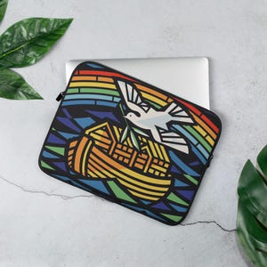 May include: A laptop sleeve with a stained glass design featuring a dove with an olive branch and a rainbow above a wooden ark. The design is on a black background.