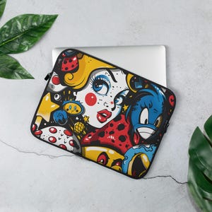 Può includere: Una custodia nera per laptop con un design a cartoni animati colorati. L'opera d'arte include il volto di una donna con occhi blu, labbra rosse e capelli gialli, insieme ad altri personaggi dei cartoni animati. La custodia è realizzata in un materiale resistente e ha una chiusura a cerniera.