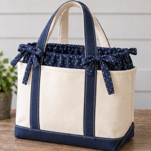 May include: A cream-colored canvas tote bag featuring navy blue trim and a base. The bag includes a navy blue drawstring insert with a white polka dot pattern, finished with two decorative bows on the front, and two sturdy navy blue handles.