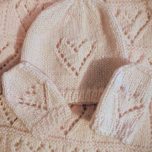 May include: A pale pink knitted baby set featuring a beanie with a heart-shaped design and matching mittens. The knitwear has a delicate, openwork pattern. The set is made from a soft, textured yarn, perfect for a newborn.
