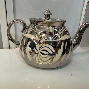 May include: A reflective silver-toned teapot featuring a rounded body and a matching lid with a pointed finial. The surface is decorated with a cream-colored floral pattern that wraps around the center, complemented by a curved handle and spout.