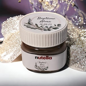 Peut inclure: Un pot de Nutella avec un couvercle et une étiquette blancs. L'étiquette et le couvercle portent l'inscription "Baptême Alma 16.09.23" avec un motif floral. Le pot est rempli d'une pâte à tartiner brun foncé.