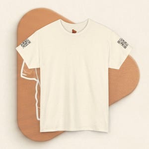 Op de afbeelding: Een crèmekleurig T-shirt met korte mouwen en een subtiele gespikkelde textuur. Het shirt is voorzien van een kleine, zwarte QR-code op de buitenrand van beide mouwen. Het kledingstuk heeft een ronde hals en een egale, lichte uitstraling.