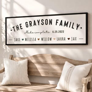 May include: A rectangular wall sign with a black frame and white background featuring the text THE GRAYSON FAMILY, Made complete 6.25.2022, and a list of names including Jake, Melissa, Willow, Laura, and Jax separated by small colored heart icons.