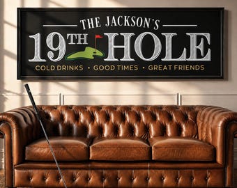 May include: A framed black sign with white text that reads "THE JACKSON'S 19TH HOLE" above a green golf course graphic. Below the text is "COLD DRINKS • GOOD TIMES • GREAT FRIENDS." A brown leather sofa and golf club are in the foreground.