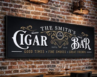 Może przedstawiać: Prostokątny czarny szyld z białym i złotym napisem THE SMITH'S CIGAR BAR, ESTD. 2018 oraz GOOD TIMES, FINE SMOKES, GREAT FRIENDS. Szyld posiada ozdobne złote zawijasy oraz małe ilustracje cygar, umieszczony na ścianie z cegły.