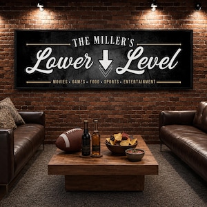 May include: A rectangular black wall sign with white text reading THE MILLER'S Lower Level, featuring a downward arrow and the words MOVIES, GAMES, FOOD, SPORTS, ENTERTAINMENT. The sign has a thin gold border and is mounted on a brick wall.