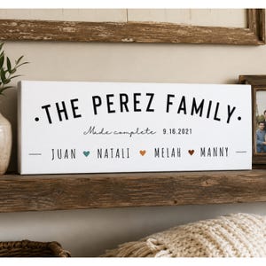 May include: A rectangular white canvas sign featuring the black text THE PEREZ FAMILY, followed by the script text Made complete and the date 9.16.2021. Below, the names JUAN, NATALI, MELAH, and MANNY are separated by small colored heart icons.
