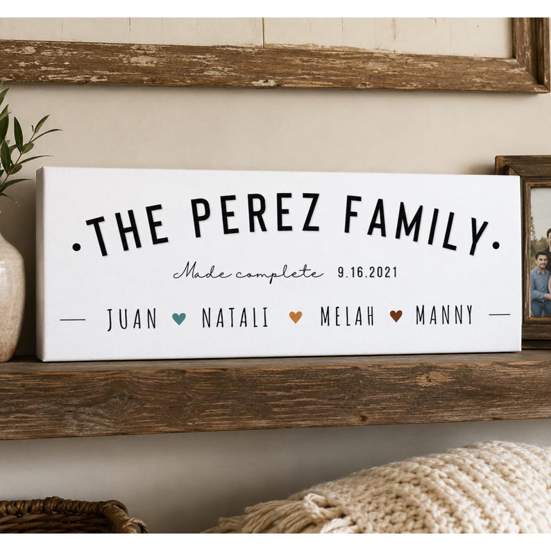 May include: A rectangular white canvas sign featuring the black text THE PEREZ FAMILY, followed by the script text Made complete and the date 9.16.2021. Below, the names JUAN, NATALI, MELAH, and MANNY are separated by small colored heart icons.