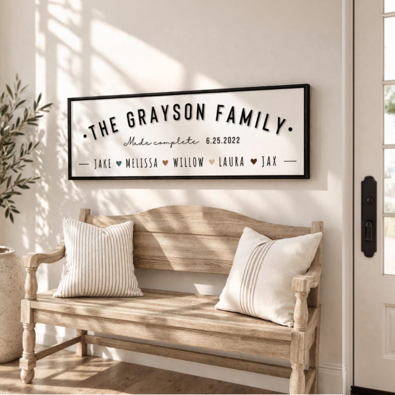 Puede incluir: Un letrero rectangular con marco negro con el texto "THE GRAYSON FAMILY" en letras negras. Debajo, se lee "Made complete 6.25.2022" y enumera nombres. Debajo, un banco de madera con dos cojines a rayas.
